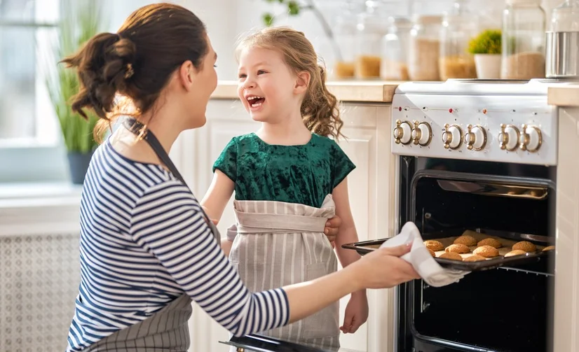 A mother using a tea towel to pull a tray of cookies out of a freestanding oven while her infant daughter smiles next to her.