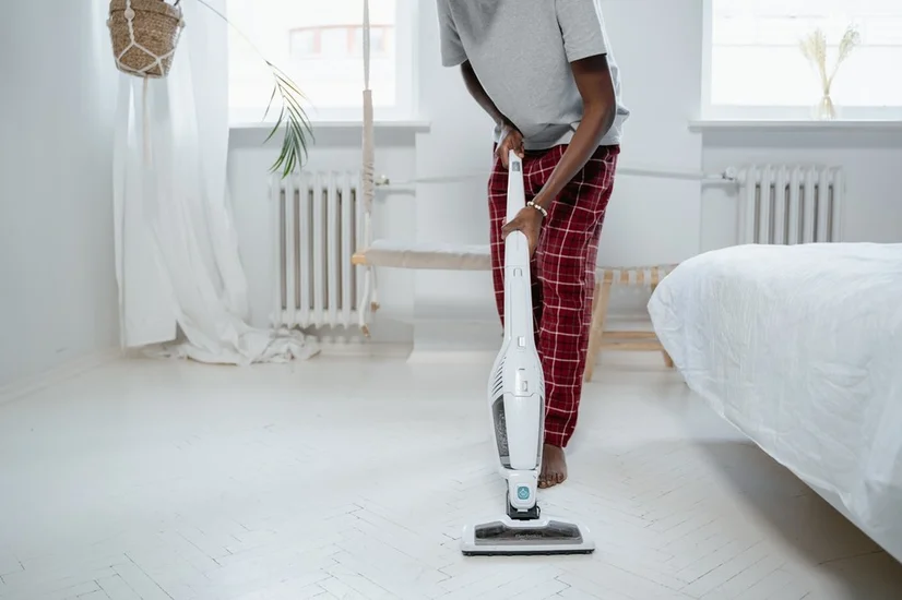 A man using a white stick vacuum to clean his bedroom floor.