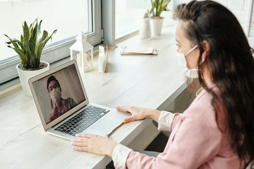 A woman wearing a surgical mask attending a telehealth appointment over video call.