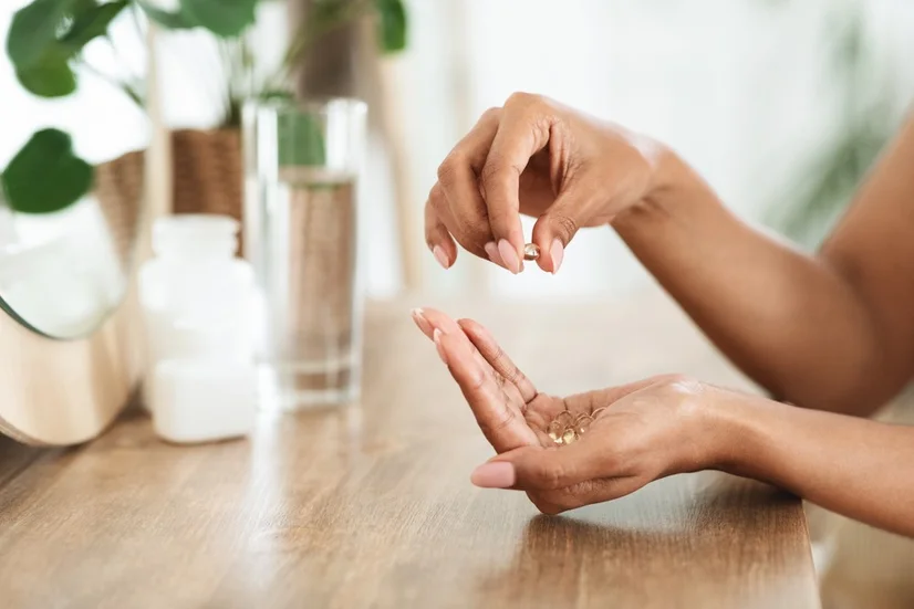 A woman holding clear vitamins in her hand. There's a glass of water in the background.