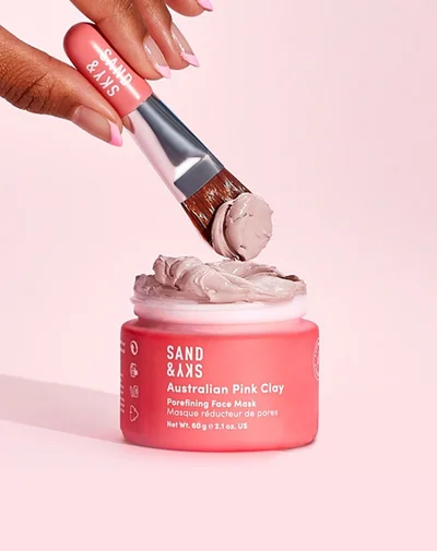 Someone using a brush to scoop some of the Sanad & Skin Brilliant Skin Pink Clay Face Mask out of its tub.