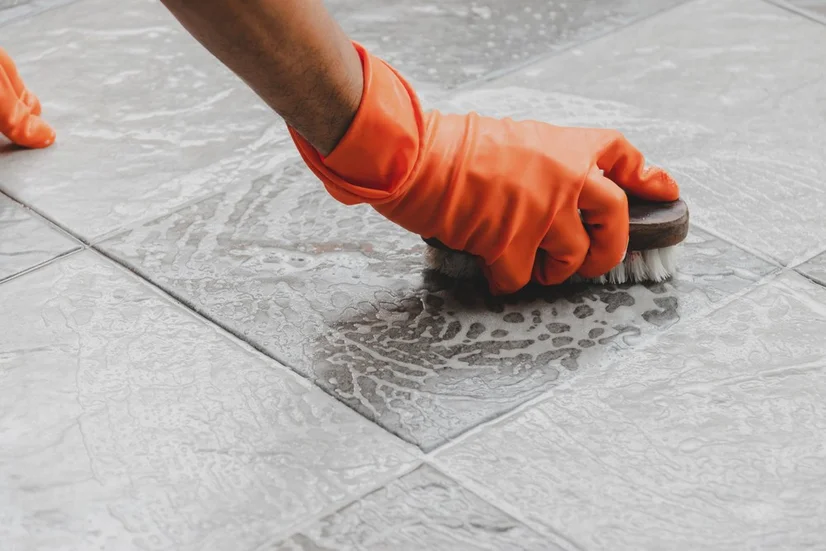 A hand wearing an orange rubber glove scrubbing grey bathroom tiles with soapy water and a brush.