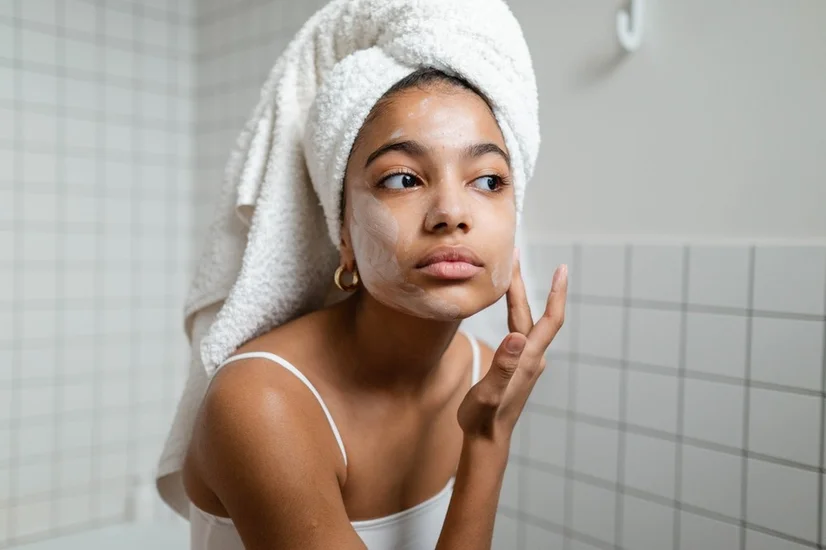 Woman with her hair wrapped in a towel applying a creamy face mask to her skin