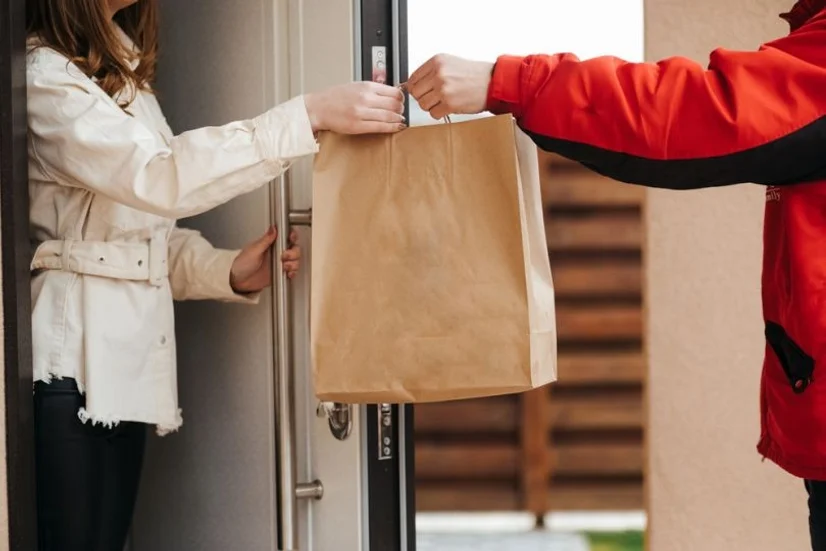 Food delivery worker handing a large brown paper bag to a woman at her front door.