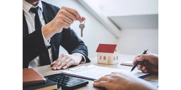A man at a desk handing over a house key to someone filling out paperwork. There's also a model of a house on the desk.