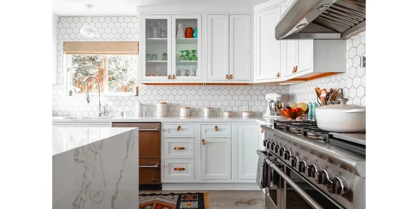A brightly lit kitchen with a marble benchtop, tiled walls and white cabinets.