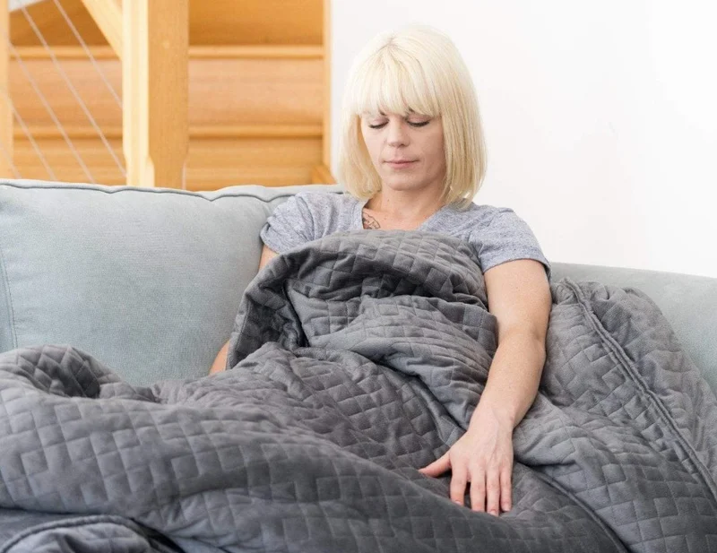 A blonde woman sitting on the couch with a grey Calming Blanket Weighted Blanket on top of her.