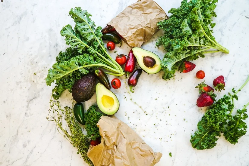 A flat lay of fresh vegetables (including kale, avocados, tomatoes, and chillis) coming out of two paper bags.