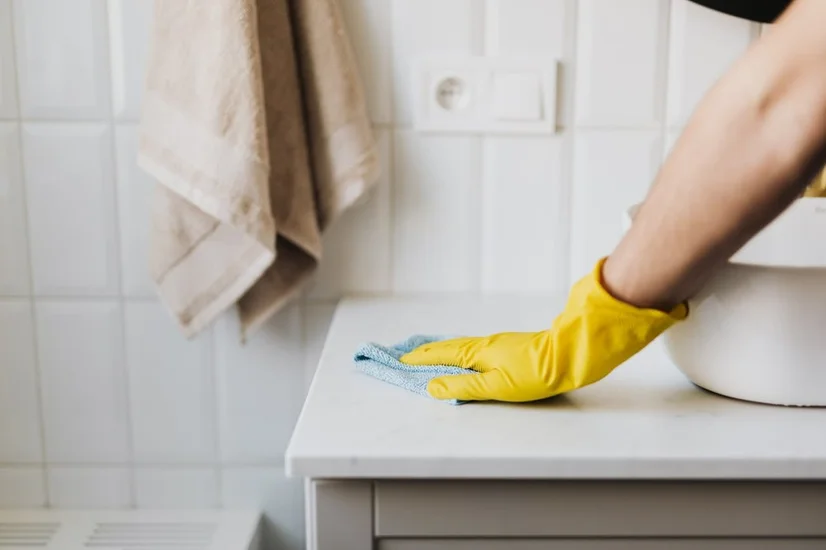 A cleaner wearing a yellow rubber glove and cleaning a bathroom sink with a cloth.
