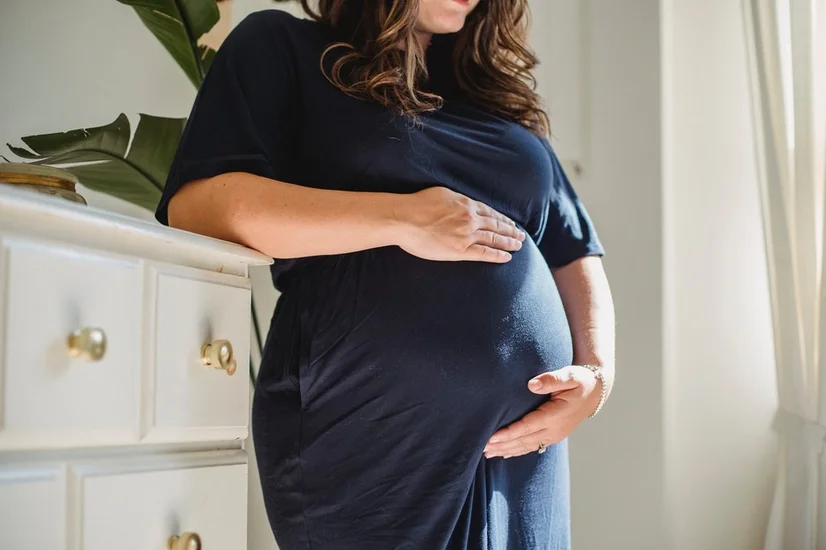 Pregnant woman in blue dress leaning against a white dresser and holding her belly