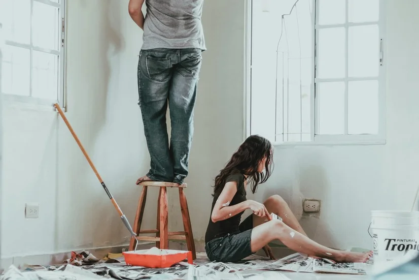 A man and woman using a roller and brush to paint a room with windows white.