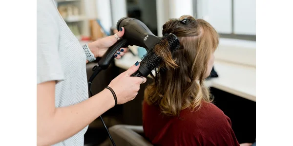 A woman in a salon having her hair dried and styled with a hair dryer and round brush.
