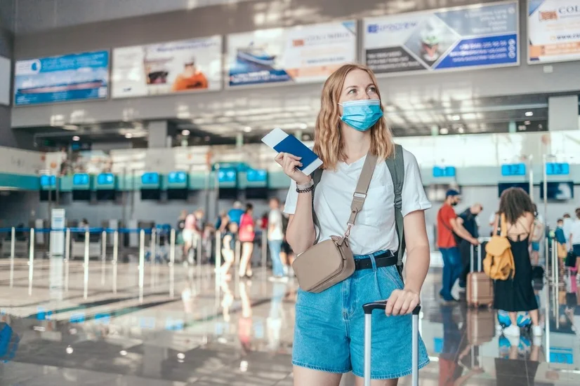 A young, blonde woman wearing a mask standing in an airport terminal and looking to the side. She's holding her passport and boarding pass and is wearing a backpack, purse, and has her hand on the handle of her suitcase.