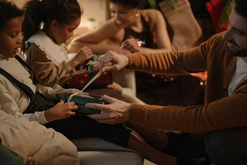 A man helping a young boy open a Christmas gift in a dimly lit room surrounded by family.