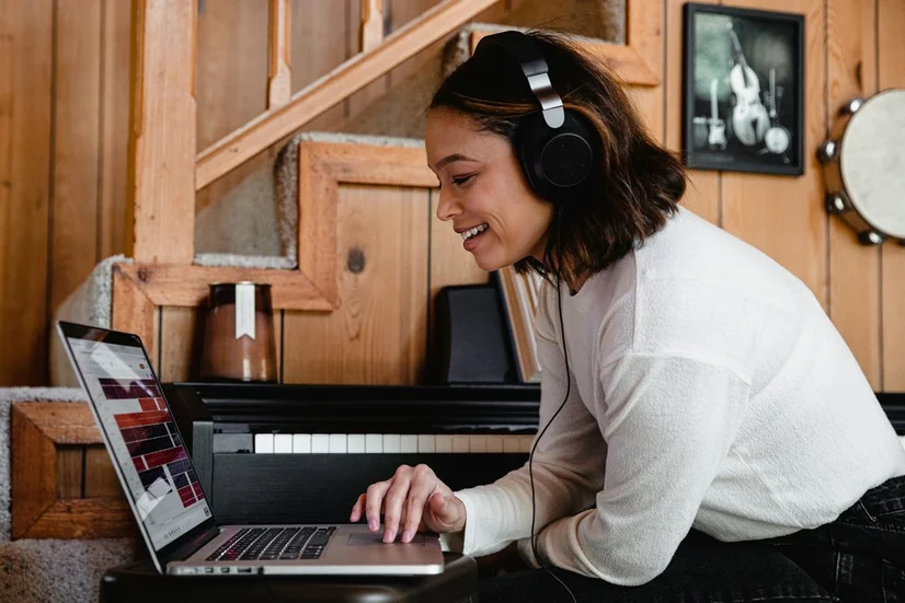 A woman wearing over-ear headphones while using a laptop. There is a piano beside her.