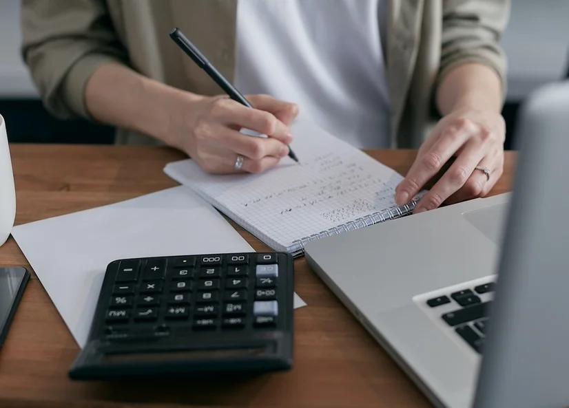 Someone making calculations on a notepad at a desk, with a calculator and laptop in front of them.