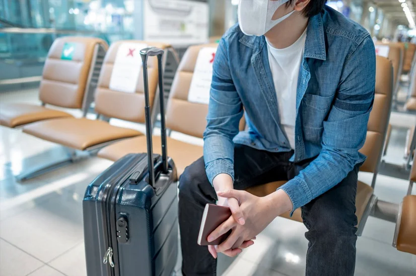 A man wearing a mask sitting down in an airport terminal. He's holding a passport and has a black suitcase next to him.