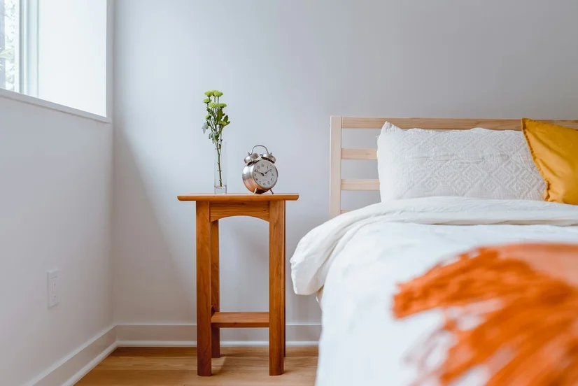 A bed with white linen in a brightly lit bedroom with a wooden bedside table next to it.