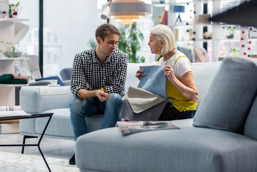 A female sales assistant showing a male customer fabric swatches while sitting on a sofa in a furniture shop.