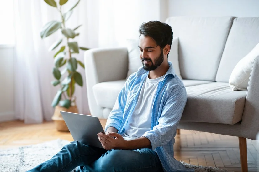 A bearded man in a blue shirt sitting on the floor in his lounge room and using a laptop.