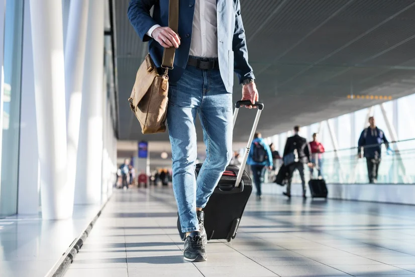 A man walking through an airport terminal wearing a satchel on his shoulder and pulling a suitcase behind him.