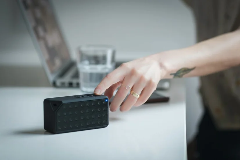 A woman turning on a black portable speaker sitting on her desk. There's a laptop and a glass of water in the background.