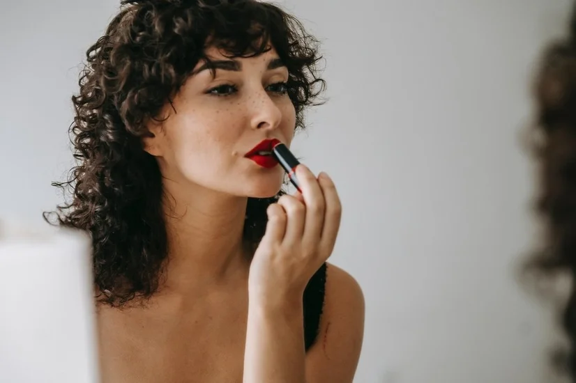 A woman with curly hair looking in the mirror and applying a red, matte lipstick.
