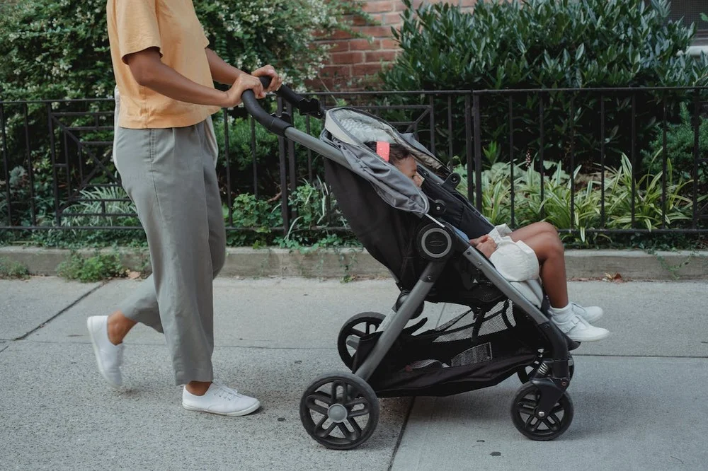 A woman pushing a four-wheeled stroller with a child inside it along a footpath.