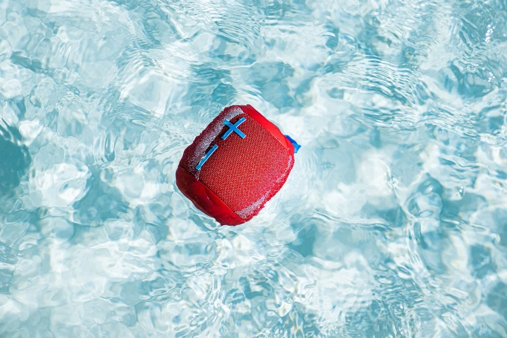 A red Logitech UE Wonderboom 2 waterproof Bluetooth speaker floating in a blue outdoor swimming pool.